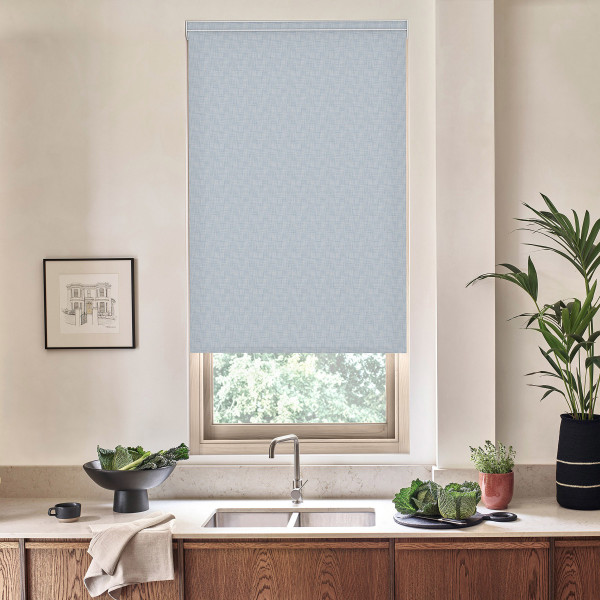 Light-blue roller blind covers most of a kitchen window, blocking light; stainless faucet and double sink sit beneath on a marble countertop, with bowls of greens, potted plants and a framed sketch nearby.