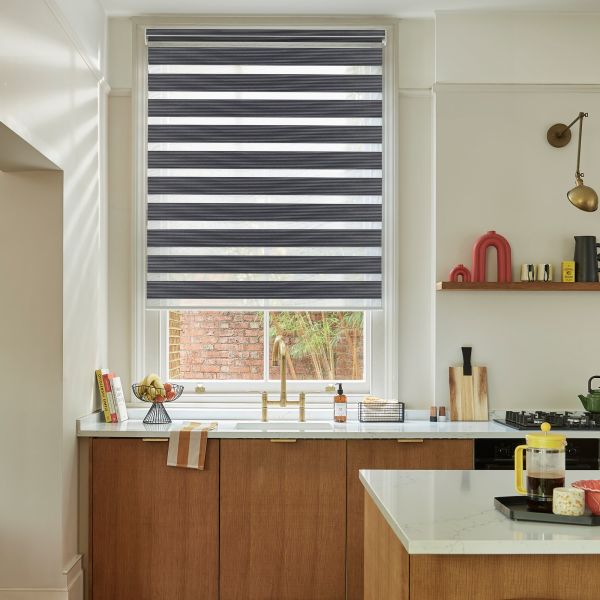 Striped dual-layer roller blinds lowered, alternating opaque and sheer bands; filtering daylight over a kitchen sink with brass faucet, white counters, wooden cabinets, and a brick-walled view outside.
