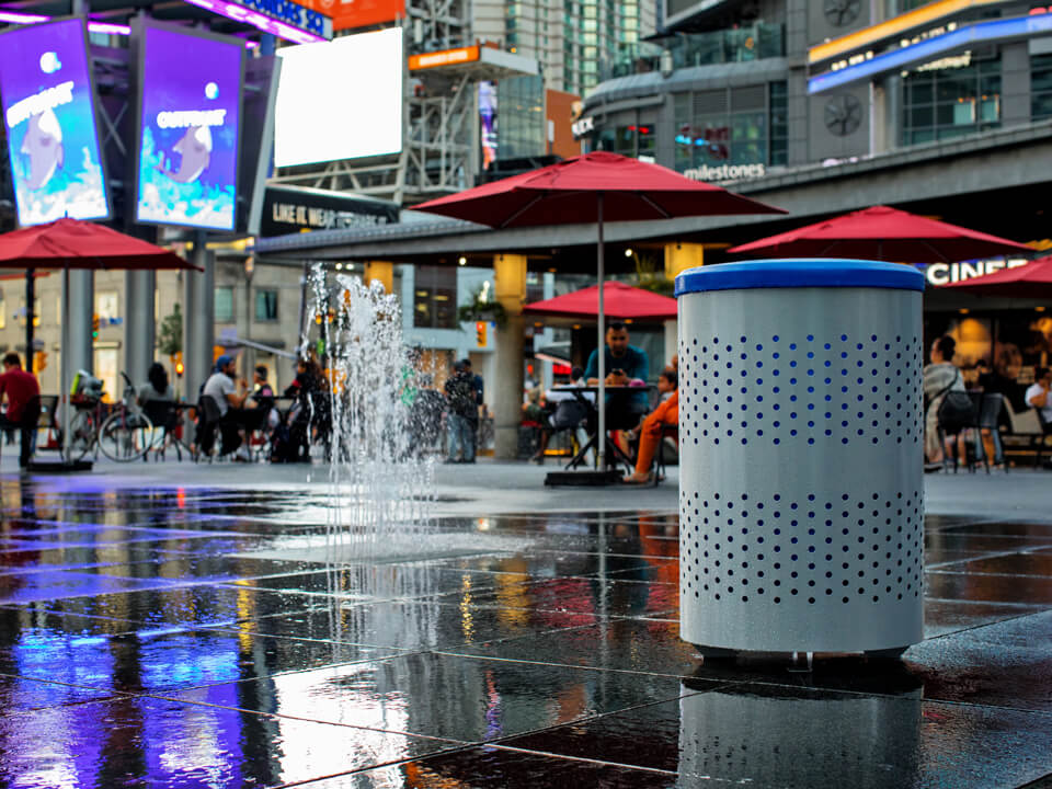silver powder coated steel recycling bin with blue plastic lid sitting at a busy downtown patio area