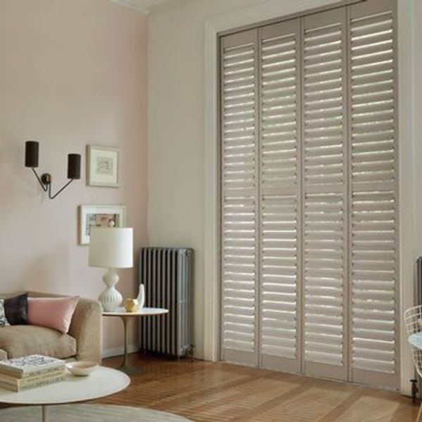 White full-height plantation shutters covering tall French doors, slatted louvers tilted to filter daylight; in a softly lit living room with pale walls, sofa, lamp, radiator, wooden floor.