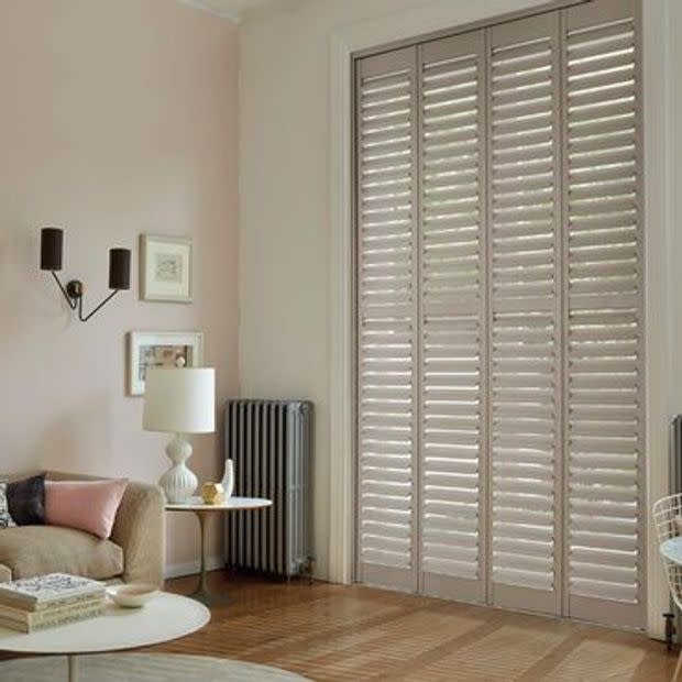 White full-height plantation shutters covering tall French doors, slatted louvers tilted to filter daylight; in a softly lit living room with pale walls, sofa, lamp, radiator, wooden floor.
