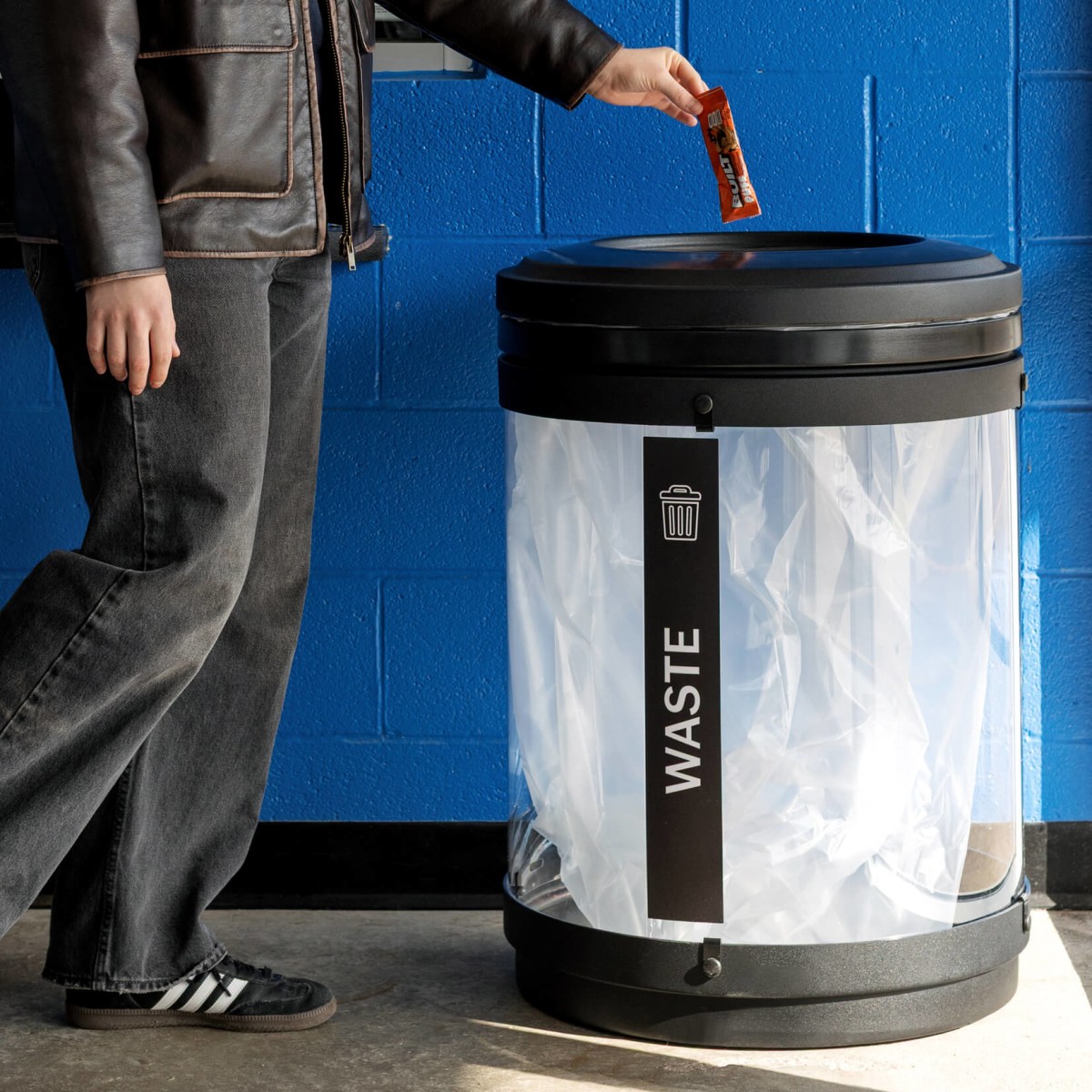 black and clear plastic waste bin indoors at a hockey arena with a person dropping waste inside