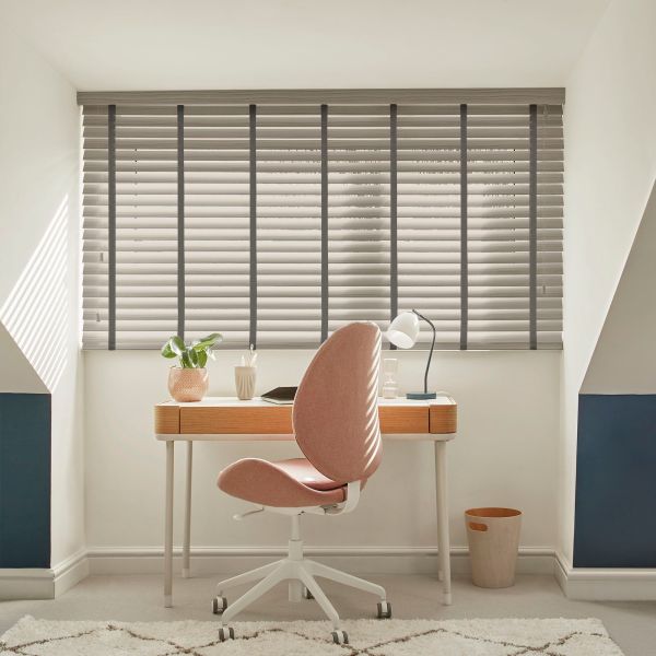 Light-gray horizontal Venetian blinds with dark vertical tapes, lowered and slats slightly tilted, filtering soft daylight into a compact attic home office with wooden desk and pink swivel chair.