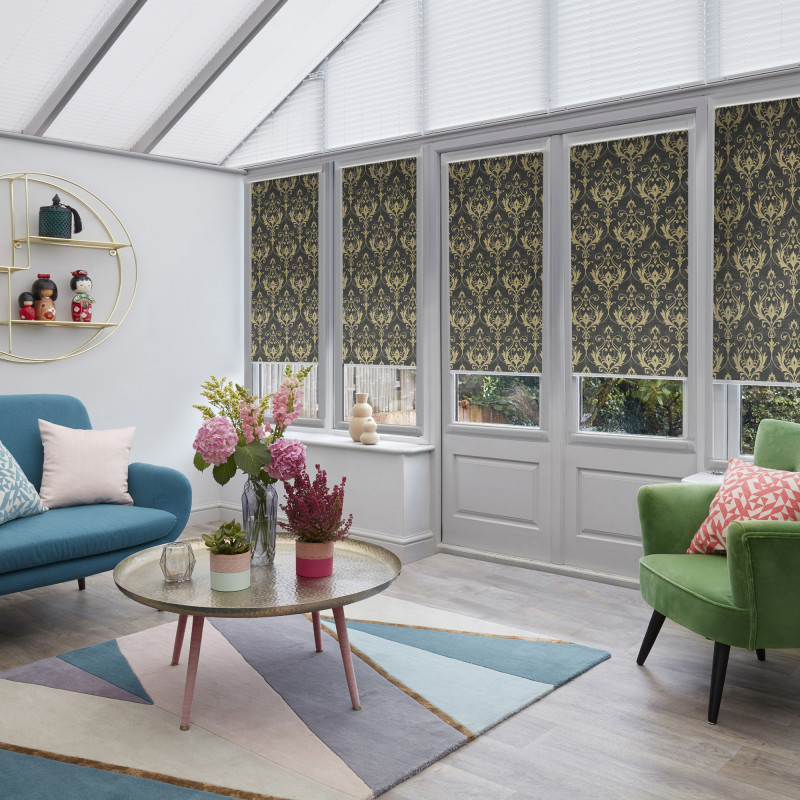 Decorative dark roller blinds with gold damask pattern, lowered over tall conservatory windows, partially blocking light; set in a bright sunroom with glass roof, colorful sofas, coffee table and flowers.