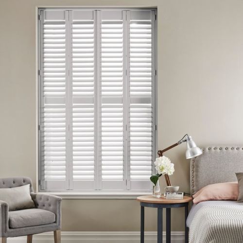White louvered plantation shutters spanning a tall window, closed with slats slightly diffusing light; bedroom setting with upholstered headboard, bedside table with lamp and flowers, and an armchair.