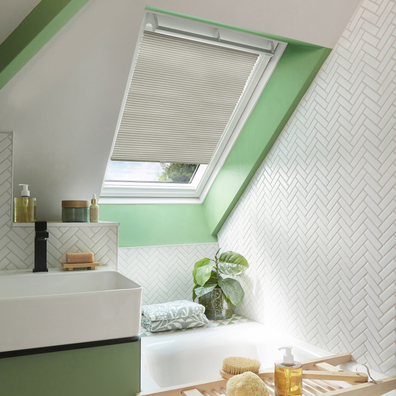Beige pleated skylight blind, half-lowered and filtering daylight, in a bright attic bathroom with green sloped window frame, white herringbone tiles, bathtub, sink, plant and toiletries.