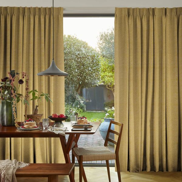 Floor-length pleated golden-yellow curtains with subtle texture, parted at center; filtering soft daylight and framing a glass door from a cozy dining area onto a sunlit, leafy backyard.