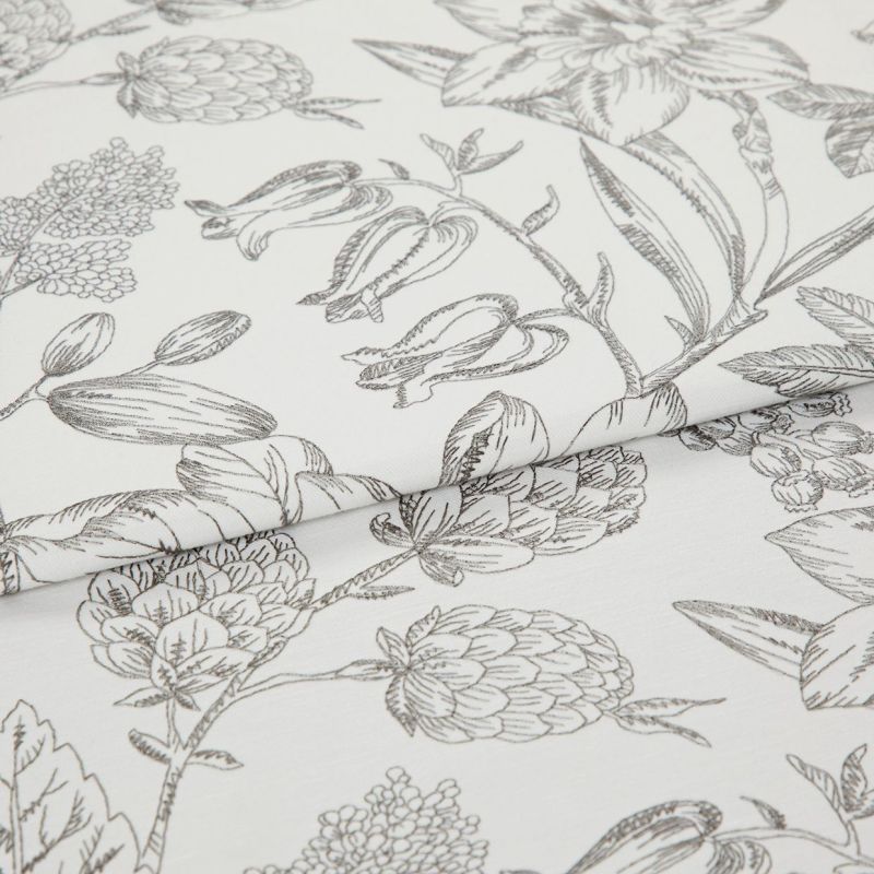 White cotton fabric with fine gray botanical sketch print, folded along the center; close-up tabletop view under soft studio lighting, emphasizing floral linework and texture.