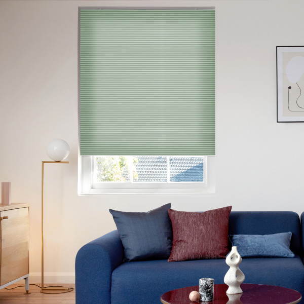 Light-green pleated cellular blind, fully lowered with horizontal folds; filtering daylight through a bottom gap, in a modern living room with a blue sofa, cushions and a floor lamp.