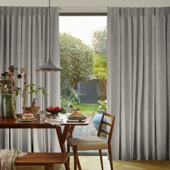 Grey floor-to-ceiling pleated curtains, parted at the center and hanging in soft folds, framing a glass door. Wooden dining table set for breakfast, pendant lamp, sunlit garden beyond.