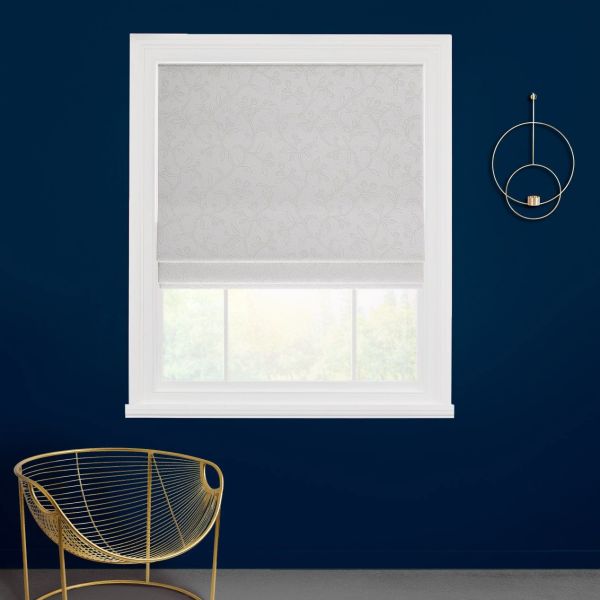 White patterned Roman shade lowered over a white-framed window, gently filtering daylight; set against a deep navy wall with a gold wire chair and circular gold wall sconce.