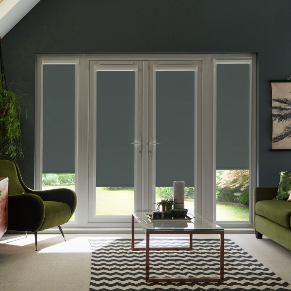French doors with dark roller blinds covering glass panels, letting narrow light strips at the bottom; glass coffee table and chevron rug between green velvet armchairs in a living room.