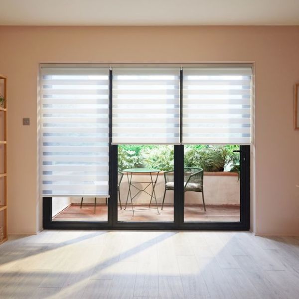Three striped dualâ€‘layer roller blinds over sliding glass doors, alternating sheer and opaque bands; partially lowered, filtering daylight onto pale woodâ€‘floored room overlooking small balcony with table, chairs and greenery.