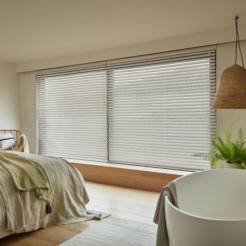 White horizontal venetian blinds across a wide window, fully closed and softly filtering daylight, in a neutral bedroom with bed draped in linen, wicker lamp, fern, and light wood floor.