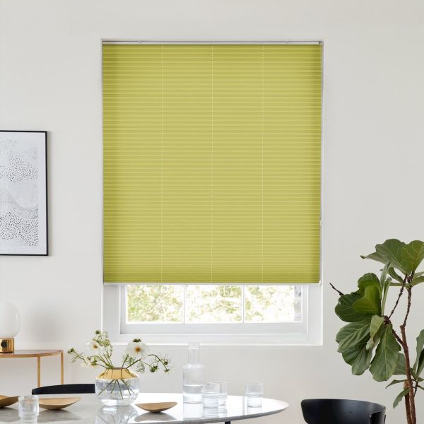 A lime-green pleated blind, fully lowered across a recessed window with vertical seams, blocking most light; modern dining area with white walls, table with glassware, flowers and a potted plant.