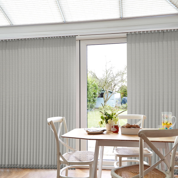 Grey fabric vertical blinds, floor-to-ceiling with slim slats and visible headrail; mostly closed but parted centrally, filtering daylight into a bright dining nook with wooden table, chairs, and a garden view.