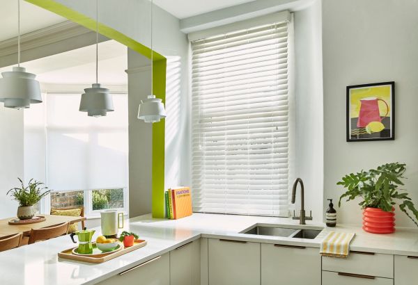 White horizontal blinds covering a tall kitchen window, closed and slightly tilted to filter sunlight and cast striped shadows; modern, bright kitchen with marble countertops, sink, pendant lights, and plants.

Text found: PANTONE