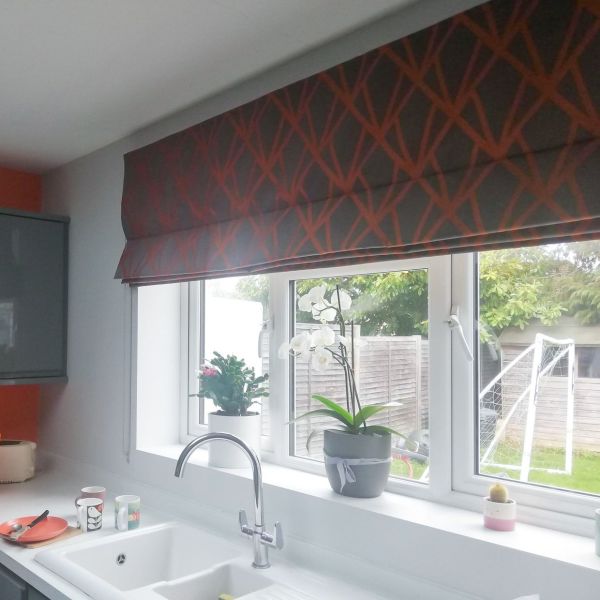 Patterned Roman blind lowered across window, folded panels with red lattice filtering light; above kitchen sink with chrome faucet, potted orchid on sill, backyard fence and small goal beyond.