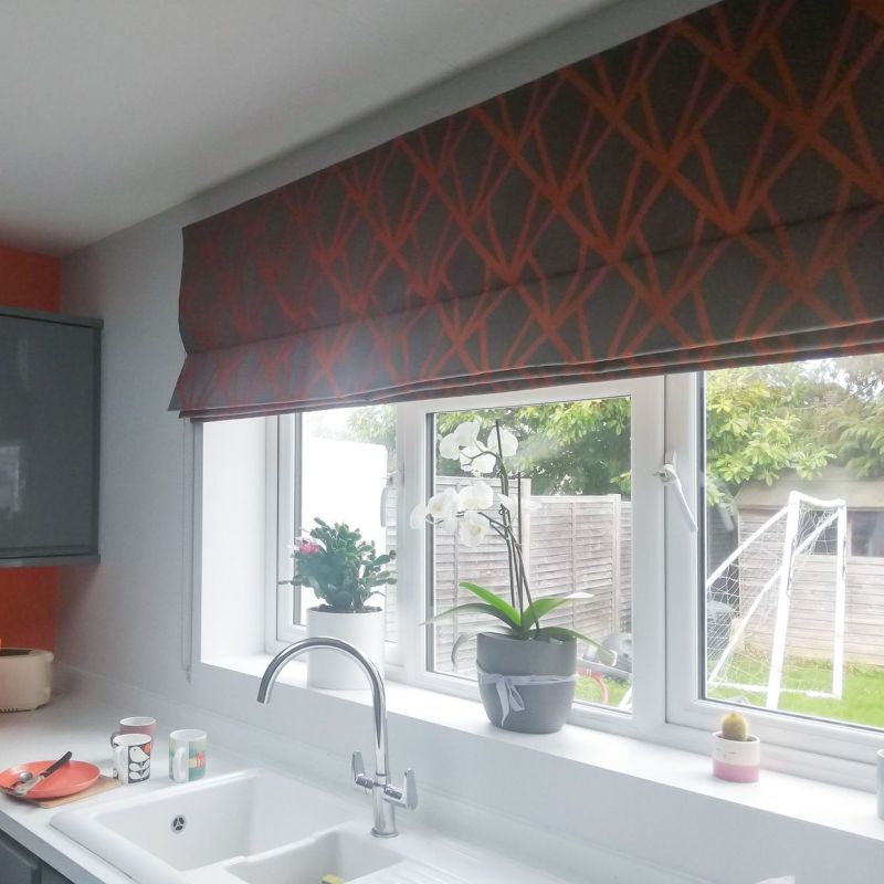 Patterned Roman blind lowered across window, folded panels with red lattice filtering light; above kitchen sink with chrome faucet, potted orchid on sill, backyard fence and small goal beyond.