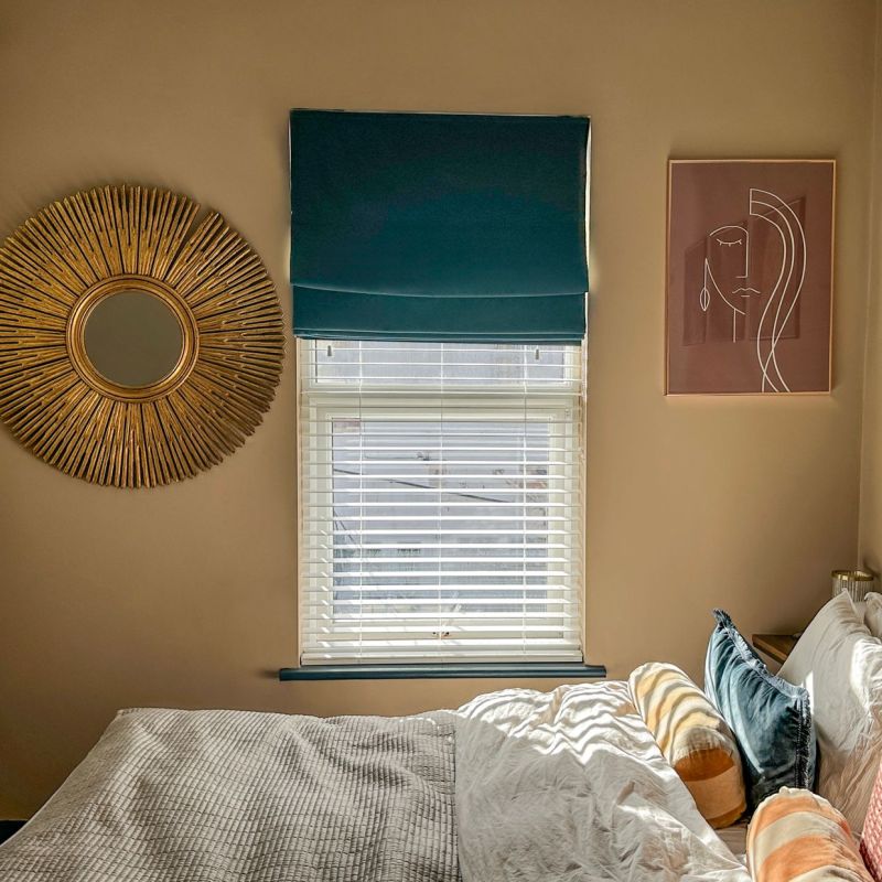 Teal Roman shade mounted above a window, partially lowered over white horizontal blinds, filtering daylight; bedroom wall with sunburst mirror left, abstract line-art print right, sunlit bed below.