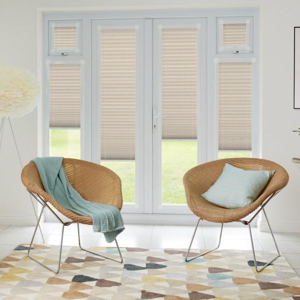 Beige pleated blinds on French doors and side panels, mostly lowered and softly filtering daylight; set in a bright living room with two rattan chairs, pastel cushions, and geometric rug.