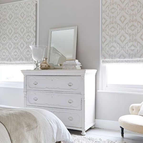 Patterned fabric Roman shades, lowered and filtering daylight through pale geometric motifs; positioned over two windows above a white distressed three-drawer dresser — neutral bedroom with mirror, vases, soft lighting.