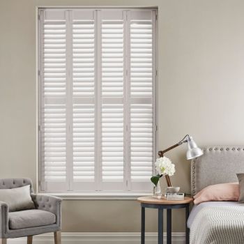 White plantation shutters spanning a tall recessed window, divided into four panels with horizontal louvers, closed and diffusing daylight; neutral bedroom setting with upholstered armchair, bedside table and lamp.