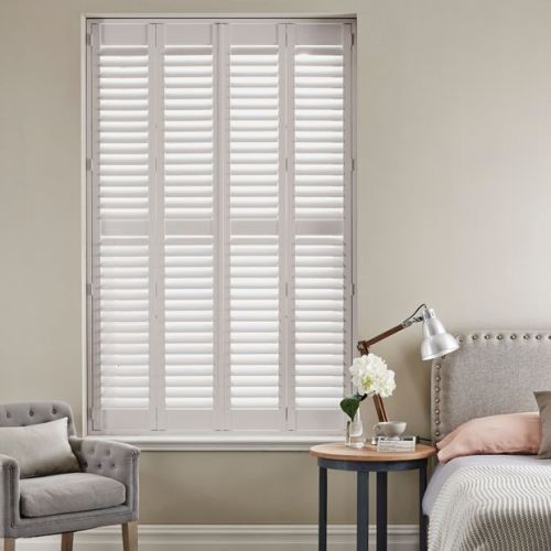 White plantation shutters spanning a tall recessed window, divided into four panels with horizontal louvers, closed and diffusing daylight; neutral bedroom setting with upholstered armchair, bedside table and lamp.