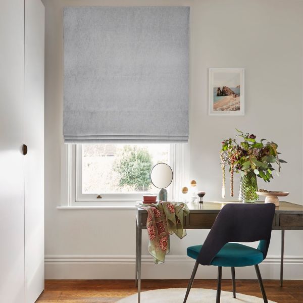 Light-grey fabric Roman blind, fully lowered, filtering soft daylight through a narrow gap; above a wooden desk in a bright office/vanity with a teal chair and a vase of flowers.