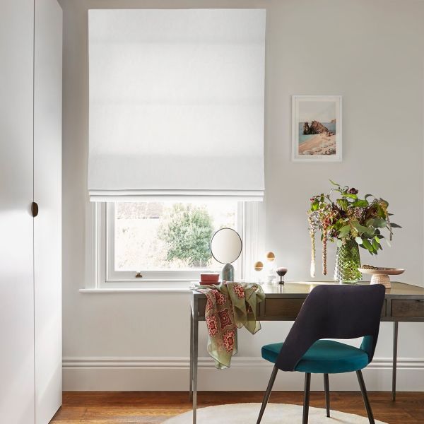 White Roman blind, lowered and slightly folded, filtering soft daylight; above a desk with teal chair, mirror and vase of flowers in a neutral-walled room, window view of greenery.