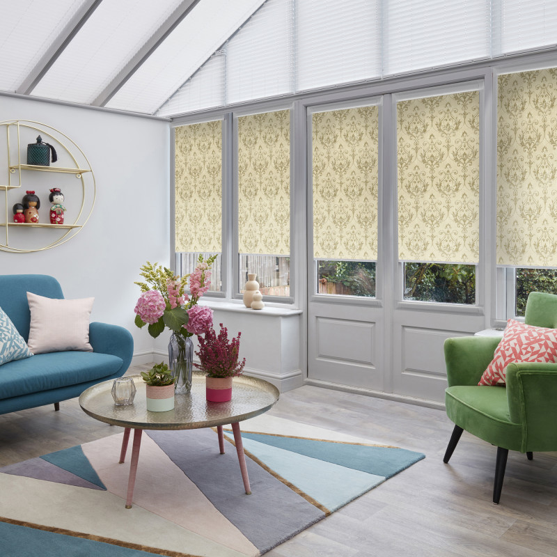 Patterned roller blinds (cream damask) cover tall rectangular windows and glass door, mostly lowered and filtering daylight; in a bright conservatory-style living room with teal sofa, green armchair, floral centerpiece.