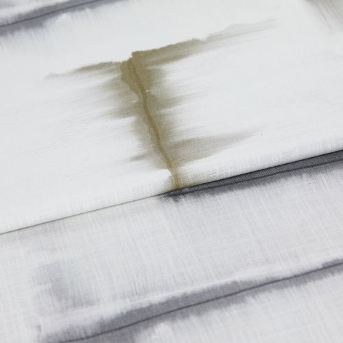Folded fabric blinds with horizontal pleats, off-white and gray-toned, marked by a vertical brown water stain; closed and stained, shown close-up in soft, diffuse indoor light.