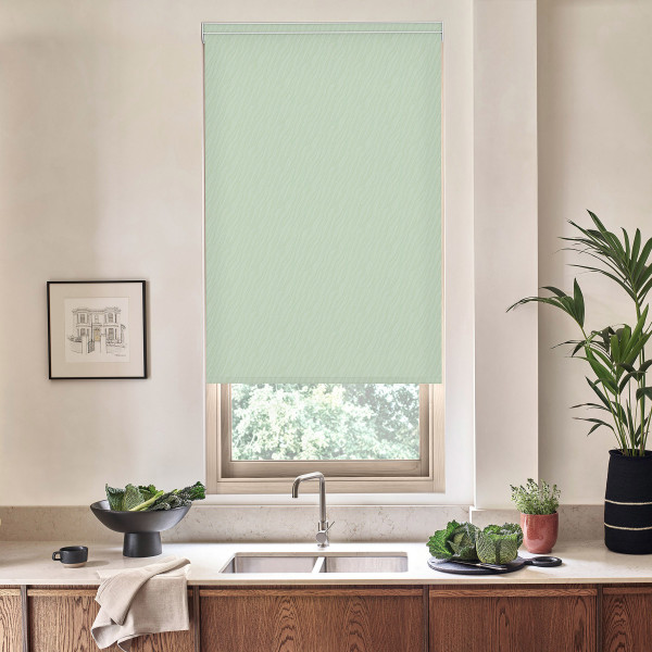 A pale green patterned roller blind covers and filters light through a kitchen window above a marble countertop with stainless sink and faucet, bowls of leafy greens and potted plants.