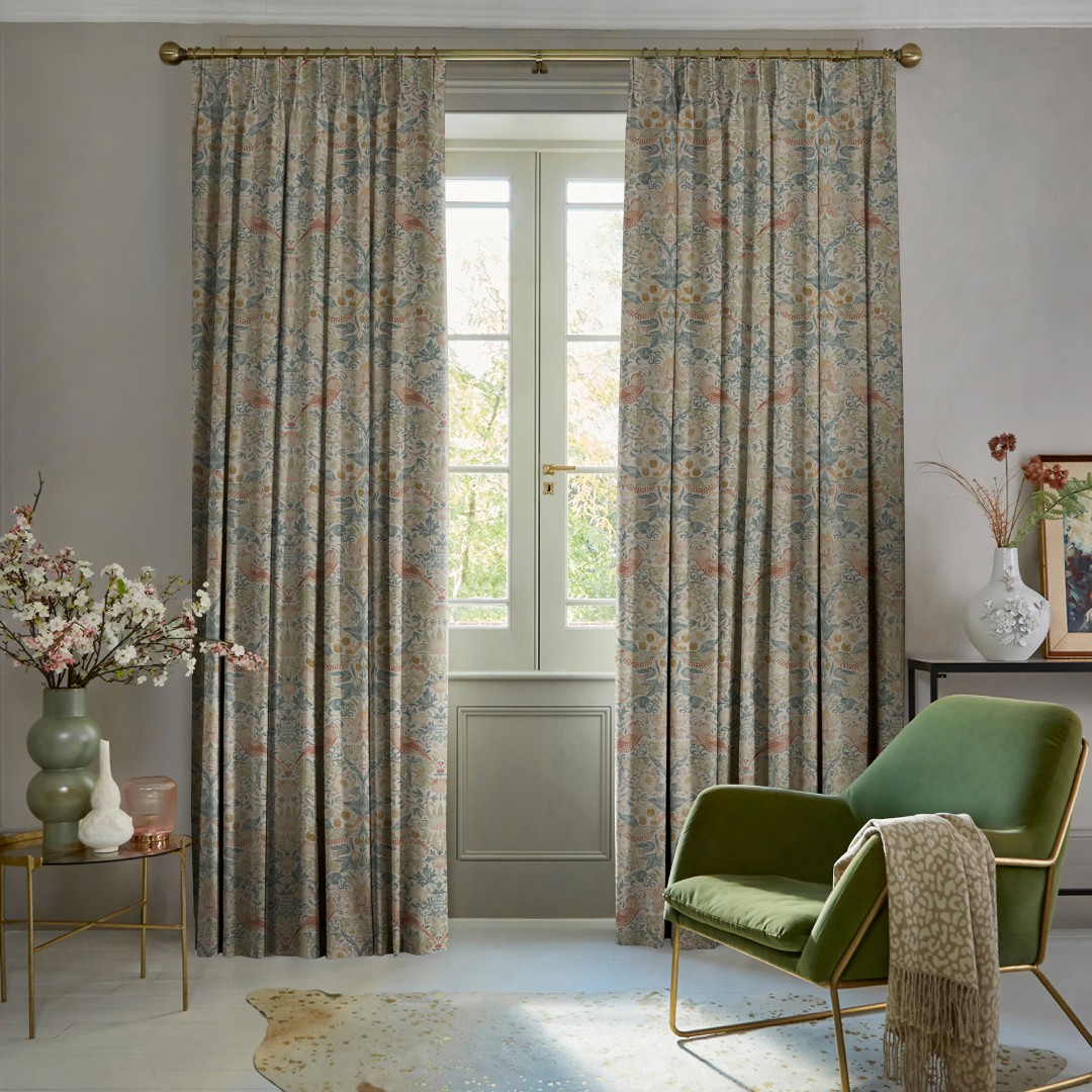 Patterned pleated full-length curtains on a brass rod, parted to reveal a French window, softly filtering daylight into a living room with green armchair, vases and a leafy garden view.
