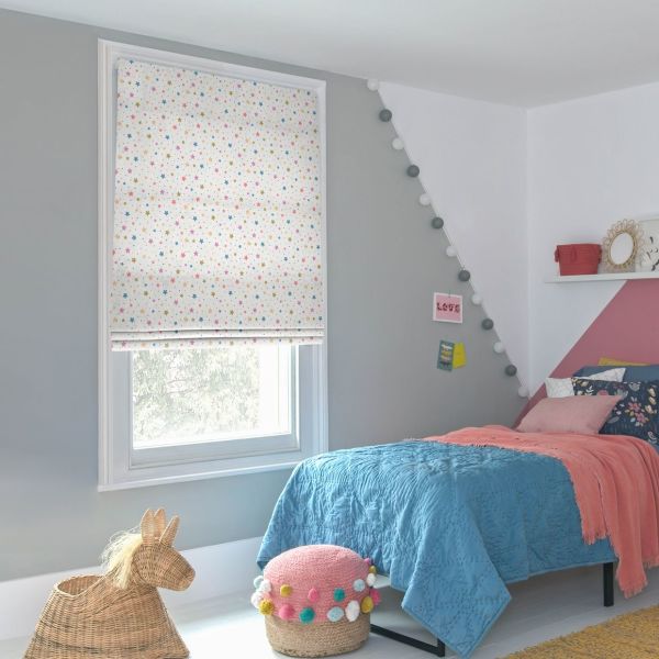 Star-patterned fabric roman blind fully lowered over the window, white with small multicolored stars, filtering soft daylight; in a girl's bedroom with gray/pink walls, blue quilt, string lights, wicker toy and pomâ€‘pom pouf. LOVE