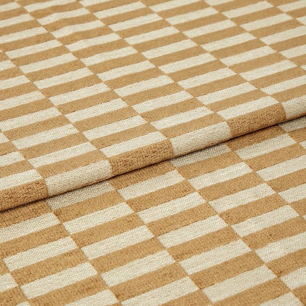 Woven beige-and-cream checkered fabric folded to create an overlapping edge, close-up studio-lit view highlighting the textured weave and repetitive rectangular pattern.