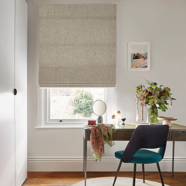 A beige textured Roman blind lowered over a sash window, gently filtering daylight; above a wooden desk with teal chair, vase of flowers and framed seaside print in an office.