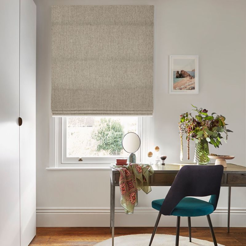 A beige textured Roman blind lowered over a sash window, gently filtering daylight; above a wooden desk with teal chair, vase of flowers and framed seaside print in an office.