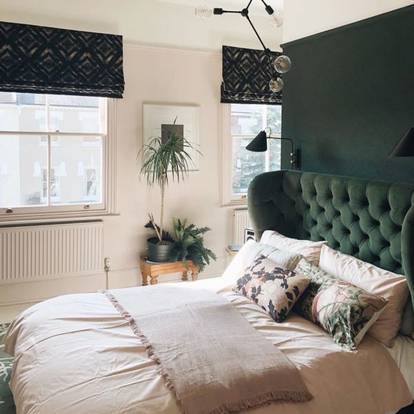 Dark patterned Roman blinds, partially lowered and folded, filtering soft daylight into a sunlit bedroom with a tufted emerald headboard, pale bedding, potted plants, wall lamps and sash windows.