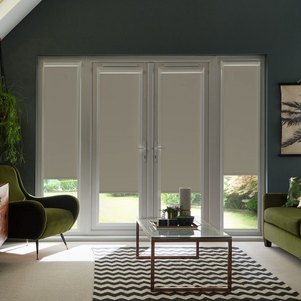 Large glass double doors with beige roller blinds partially lowered reveal a sunlit green garden, framed by dark walls, mid-century green armchairs and a glass coffee table on a chevron rug.