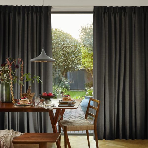 Floor-length dark gray pleated curtains, gathered to sides, pulled open at center, framing a dining table with lamp and chairs, opening onto a sunlit backyard with trees and gate.
