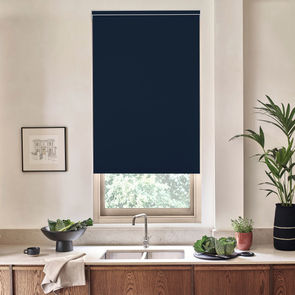 Window with a dark navy roller blind lowered above a stainless-steel sink and faucet, set in a minimalist kitchen countertop with potted plants, bowls of greens, and wood cabinets.