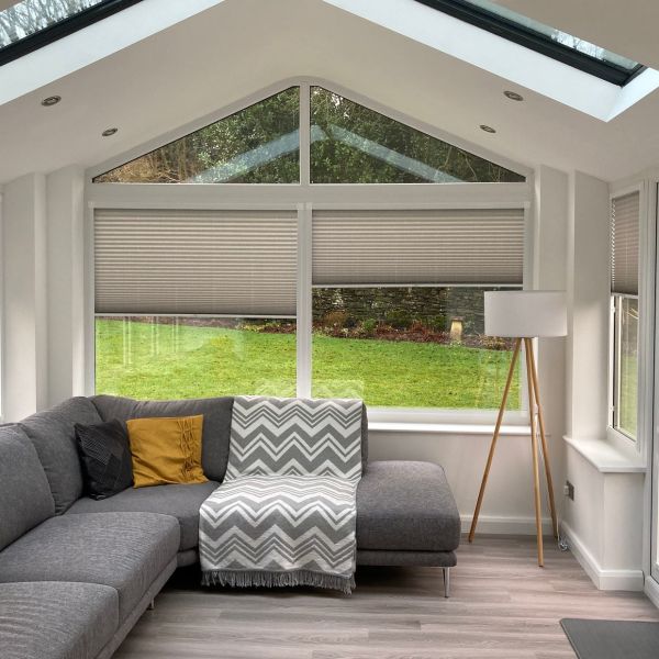 Pleated cellular blinds across a triangular-topped bay window, partially lowered to mid-height and filtering daylight; set in a bright conservatory-style sitting room with grey L-shaped sofa, floor lamp, and lawn view.