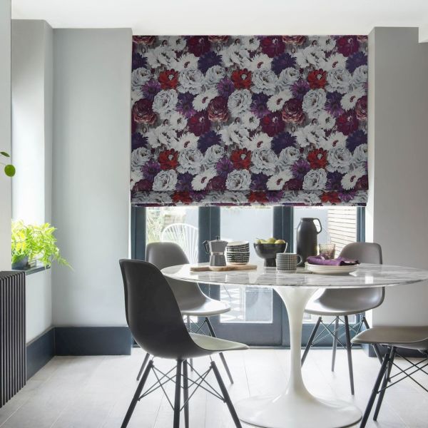 Floral Roman blind, lowered over patio doors with dense white, purple and red blooms; filtering soft daylight into a modern dining nook with a white tulip table, grey chairs, plant.