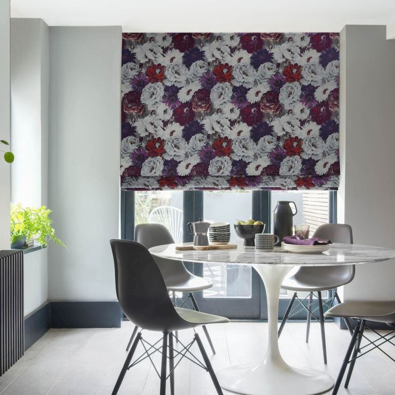 Floral Roman blind, lowered over patio doors with dense white, purple and red blooms; filtering soft daylight into a modern dining nook with a white tulip table, grey chairs, plant.