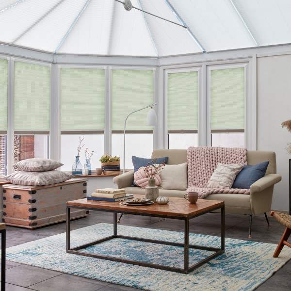Pale green pleated cellular blinds, lowered and closed, filtering soft daylight across tall conservatory windows; cozy living room with beige sofa, chunky knitted throw, wooden coffee table, and blue rug.