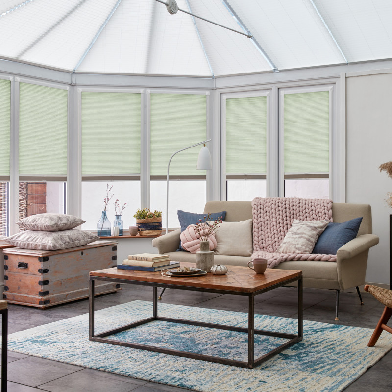 Pale green pleated cellular blinds, lowered and closed, filtering soft daylight across tall conservatory windows; cozy living room with beige sofa, chunky knitted throw, wooden coffee table, and blue rug.