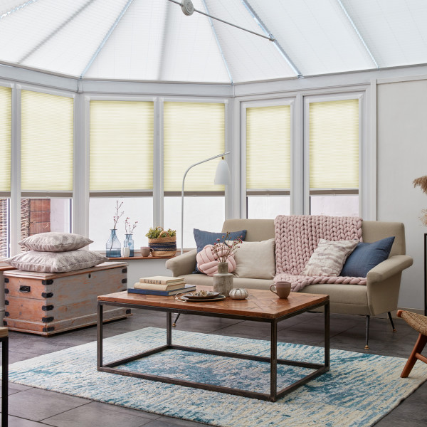 Pleated cellular blinds covering conservatory roof and bay windows, partially lowered and filtering soft daylight; in a bright, cozy sunroom with sofa, coffee table, woven rug, and minimal outside view.