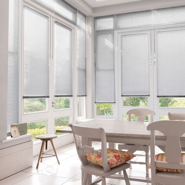 Horizontal aluminum Venetian blinds on tall bay windows, lowered to mid-height and filtering soft daylight, in a bright dining nook with a white table, chairs, cushions, tiled floor, and garden view.