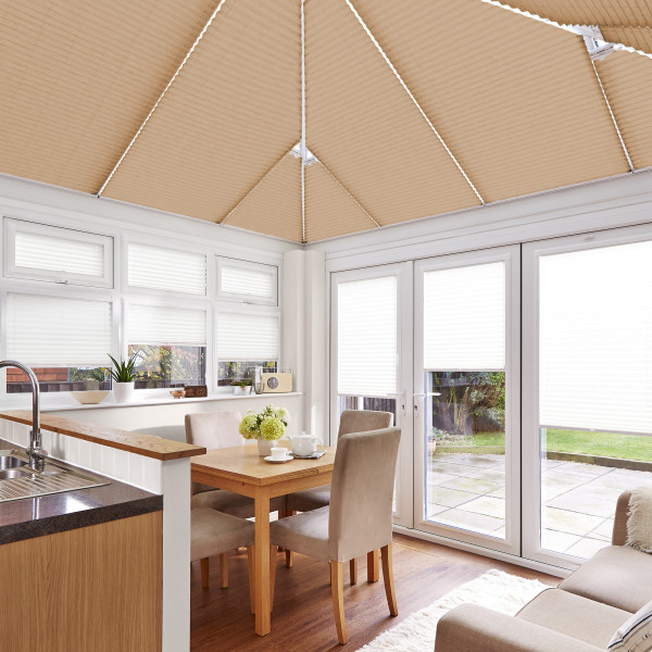 Beige pleated roof blinds and white pleated window/door blinds, fully lowered, filtering soft daylight; in a bright conservatory dining area with wooden table, upholstered chairs, sink and garden patio view.